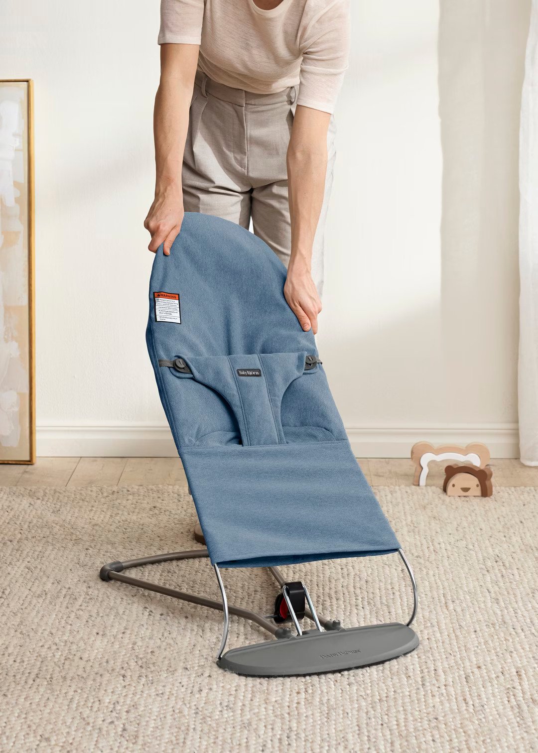 A person is adjusting a blue baby bouncer chair on a carpeted floor in a softly lit room.