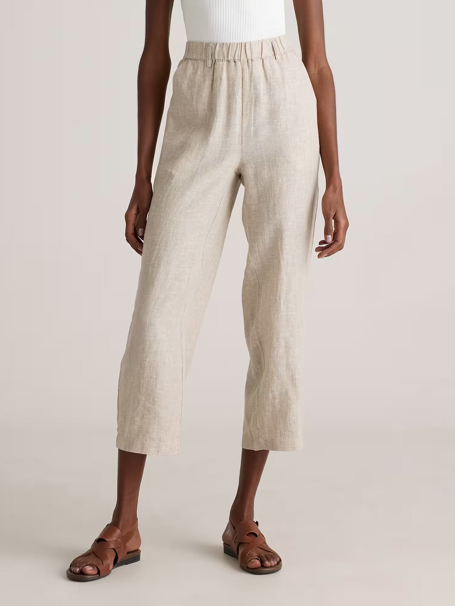 A person wearing beige, cropped, wide-leg linen pants with an elastic waistband, a white sleeveless top, and brown sandals, standing against a plain background.