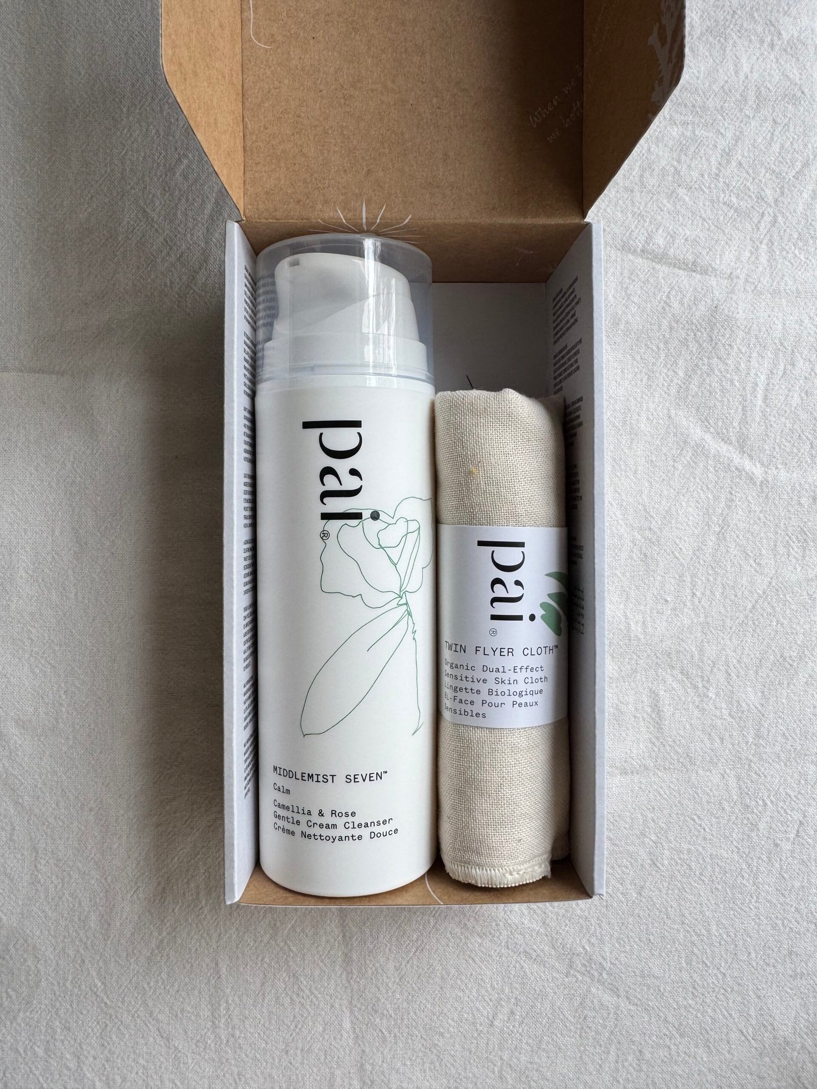 A bottle of Pai face cleanser and a towel inside a cardboard box on a white fabric surface.