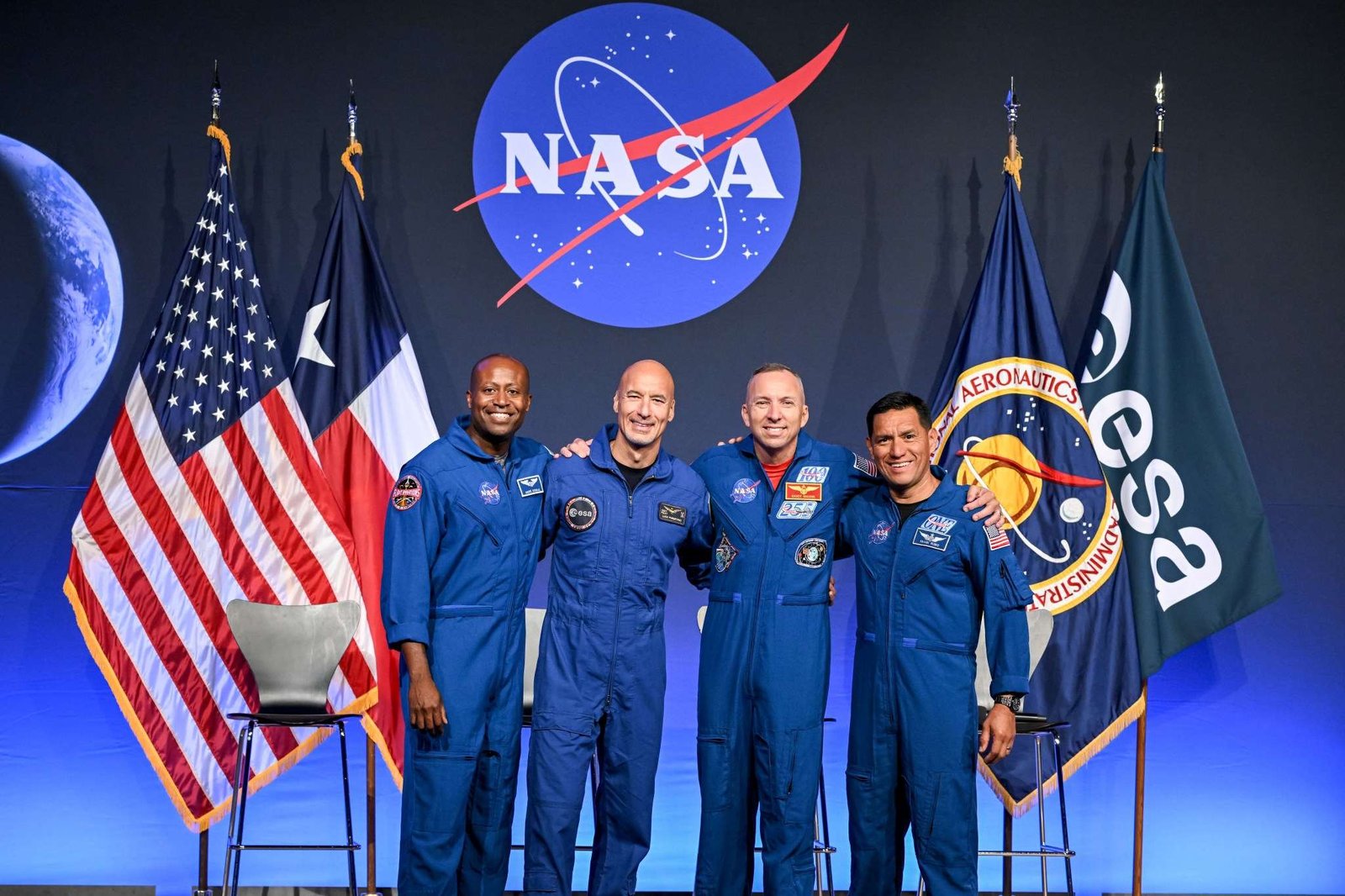 From left to right, NASA astronaut Andre Douglas, ESA (European Space Agency) astronaut Luca Parmitano, and NASA astronauts Randy Bresnik and Frank Rubio take a photo together. Their arms are linked around each other's shoulders. Behind them are the flags of the United States, Texas, NASA, and ESA.