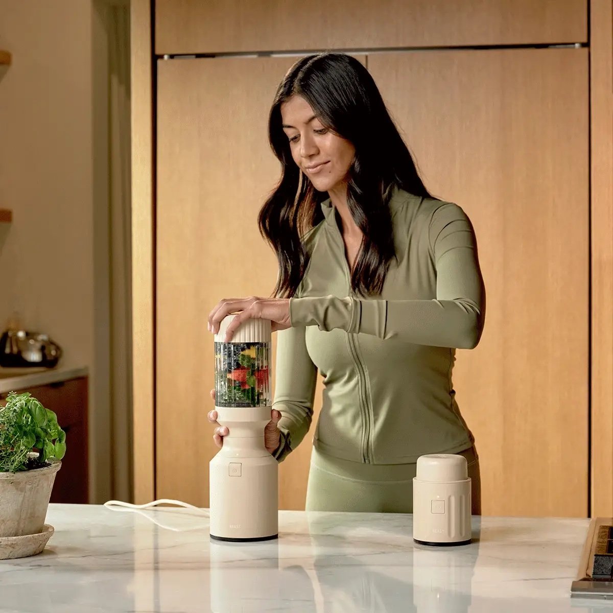 A woman in a light green outfit uses a portable blender with fruits and vegetables inside on a kitchen counter.