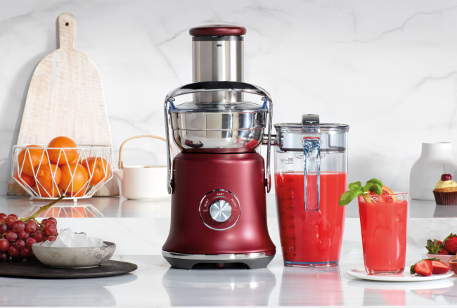 A red juicer sits on a kitchen counter with a pitcher and glass of red juice, grapes, and a bowl of oranges in the background.