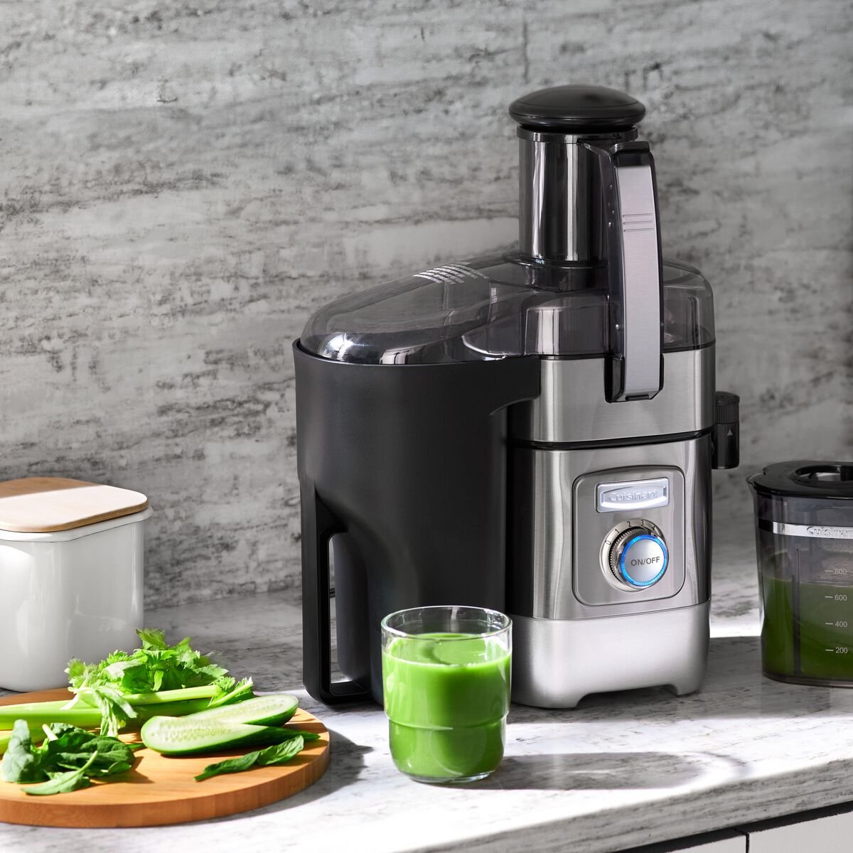 A silver electric juicer sits on a kitchen counter with a glass of green juice, leafy greens, and cucumber slices nearby.