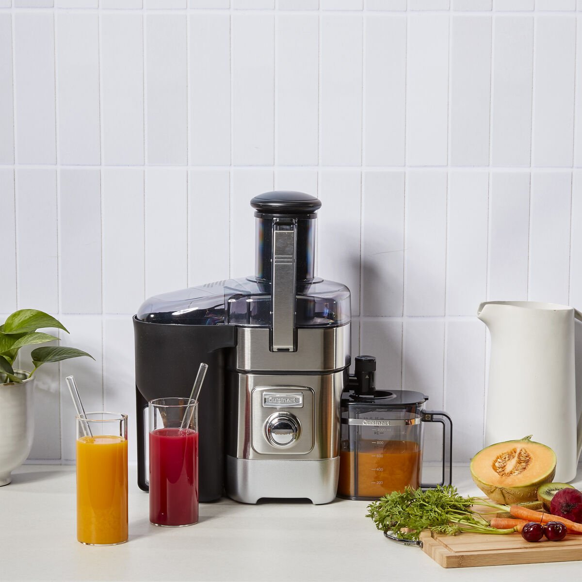 A stainless steel juicer sits on a kitchen counter with two glasses of juice, a pitcher, chopped fruit, vegetables, and a potted plant nearby.