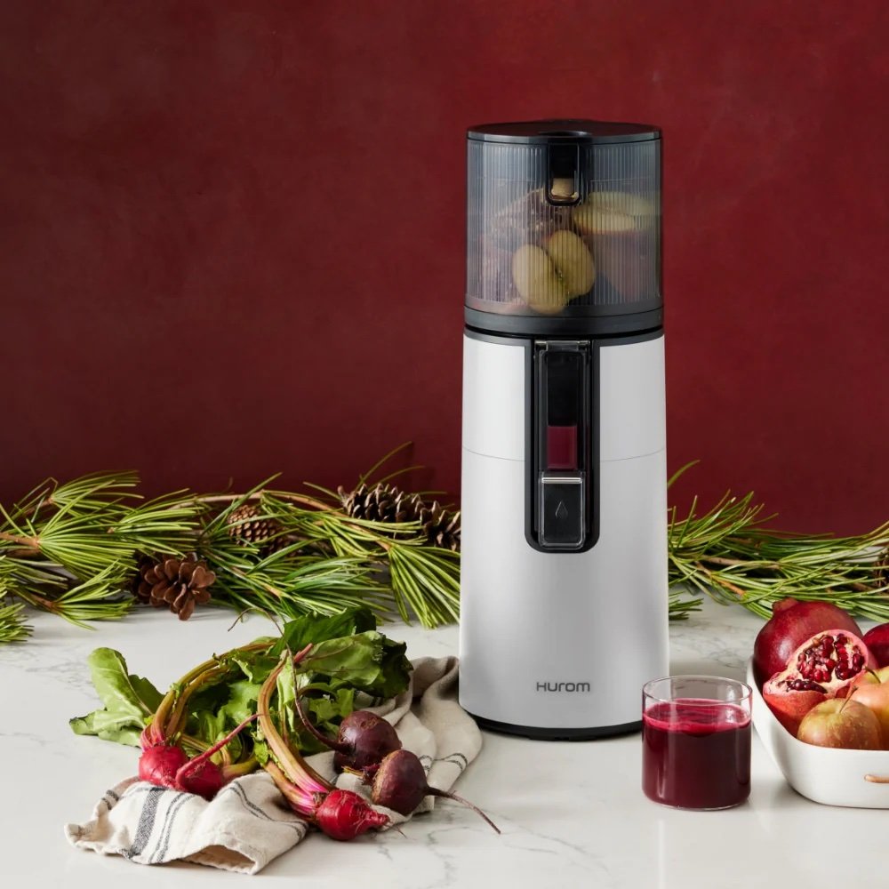 A silver Hurom juicer with fruits and vegetables inside, a glass of juice, beets, pomegranate, and kitchen towel on a marble counter with pine branches in the background.