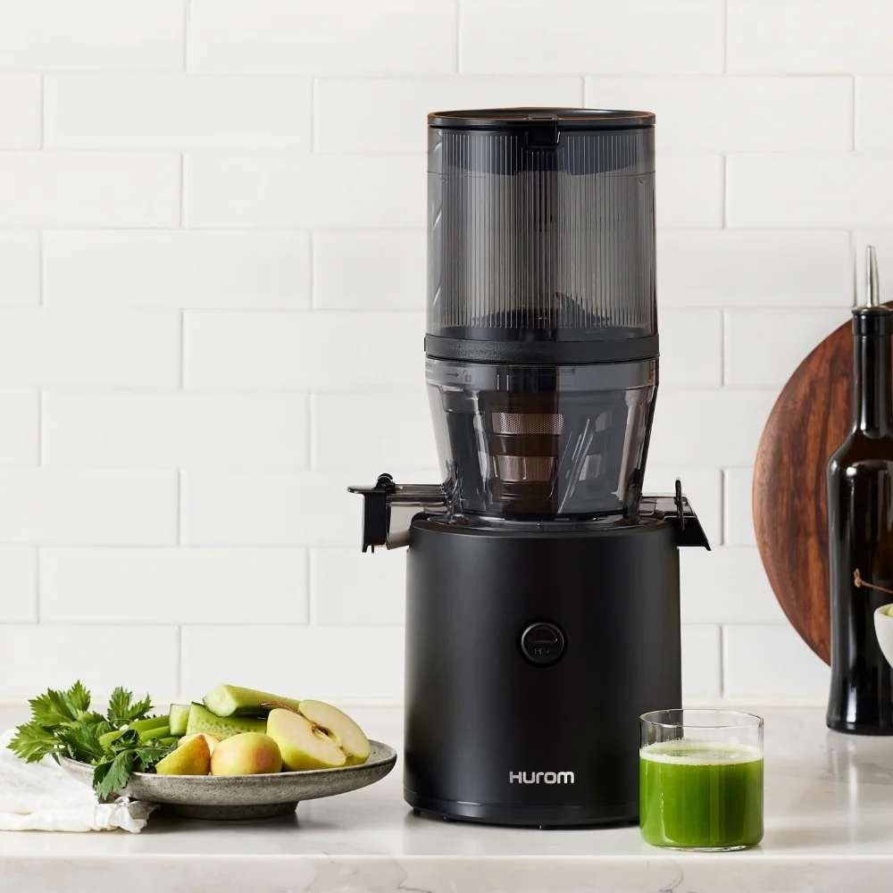 A black Hurom juicer on a kitchen counter with a plate of apples and celery, and a glass of green juice beside it.