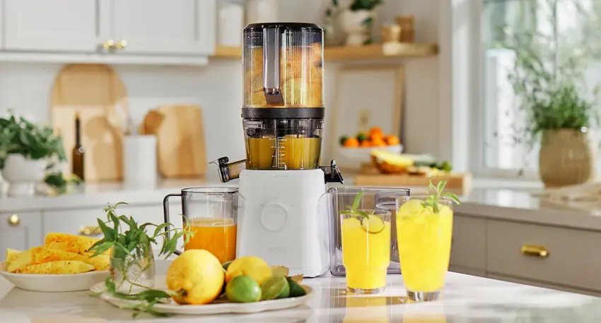 A juicer is processing fruit on a kitchen counter, with glasses of juice, whole lemons, limes, and pineapple pieces arranged nearby.