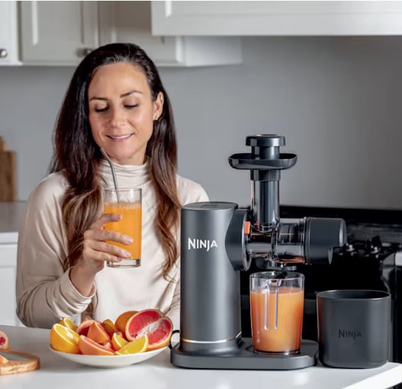 A woman sits at a kitchen counter, holding a glass of orange juice next to a Ninja juicer and a plate of sliced citrus fruits.
