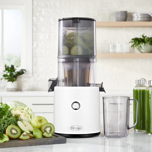 A white Omega juicer with a container of whole kiwis is on a kitchen counter, surrounded by chopped fruits, greens, and two jars of green juice.