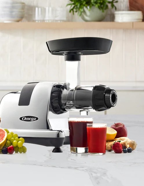 A countertop juicer with two glasses of red juice in front, surrounded by assorted fruits and ginger on a marble surface.