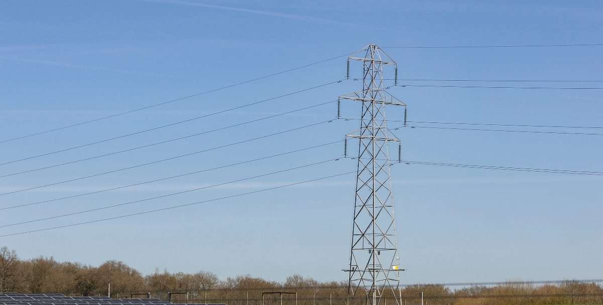 Overhead power lines and an electricity pylon, seen in profile with tree-covered ground just visible at the bottom and blue sky otherwise dominating the image
