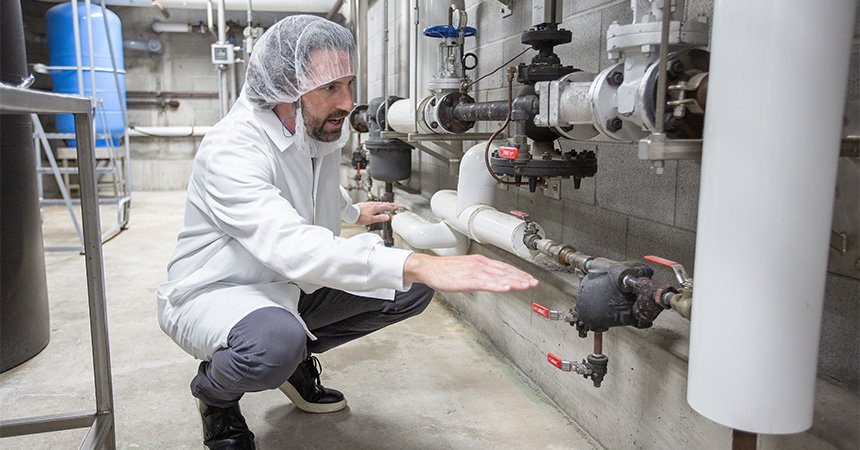 A worker in a white lab coat and hairnet crouches beside a wall-mounted system of industrial pipes and red-handled valves, gesturing toward the equipment inside a clean manufacturing facility.