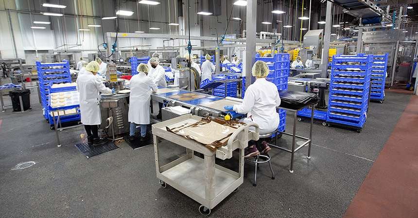 Workers in white lab coats, hairnets, and gloves process and package food at stainless steel tables inside a brightly lit industrial food production facility, with blue racks of trays and machinery in the background.