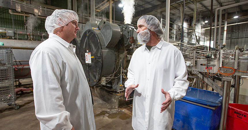 Two workers in white lab coats, hairnets, and beard covers talk inside an industrial processing facility, surrounded by large metal machinery, pipes, and steam.