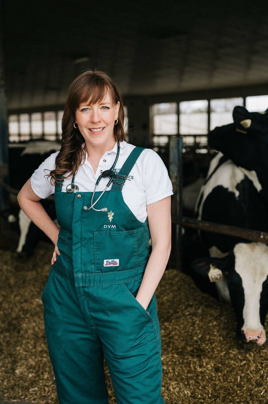Dr. Shari van de Pol, in green DVM coveralls with a stethoscope, stands in a dairy barn with Holstein cows behind her.