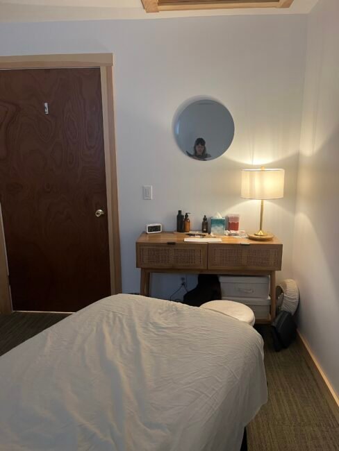 A small room with a massage table, a wooden dresser holding bottles and tissues, a lamp, and a round mirror reflecting a person.