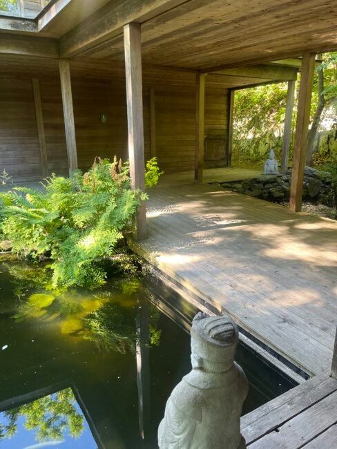 Wooden walkway with a pond, green ferns, and two stone statues, one by the water and one in the background, surrounded by trees and natural light.