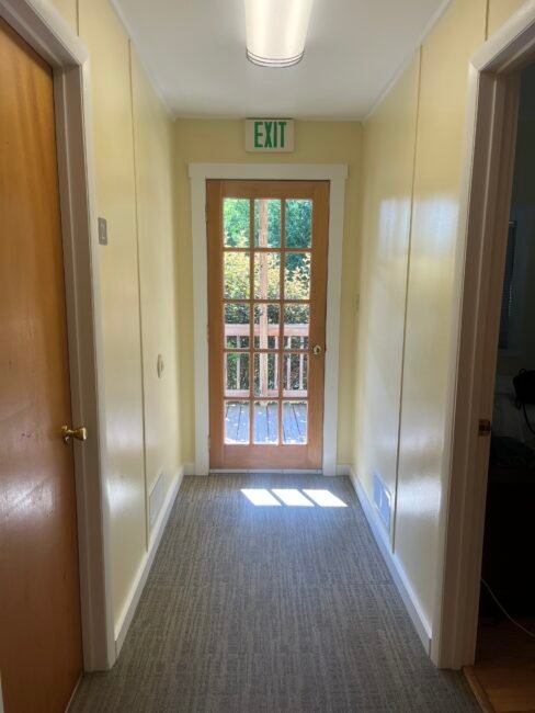 A narrow hallway with yellow walls, two closed wooden doors on each side, and a glass door at the end leading outside with an exit sign above it. Sunlight shines through the glass.