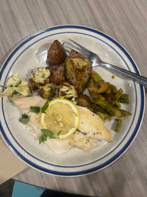 A plate with baked white fish topped with a lemon slice, roasted red potatoes, cauliflower, and green beans, with a fork on the side.