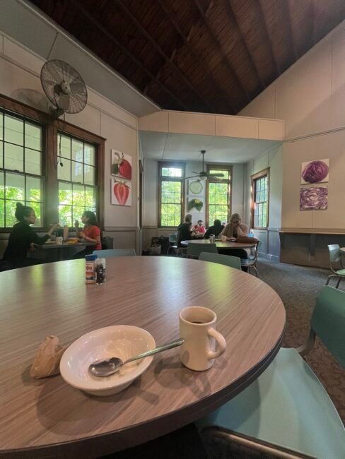 A mostly empty dining room with several people seated at tables, empty dishes on the closest table, and large windows letting in natural light.