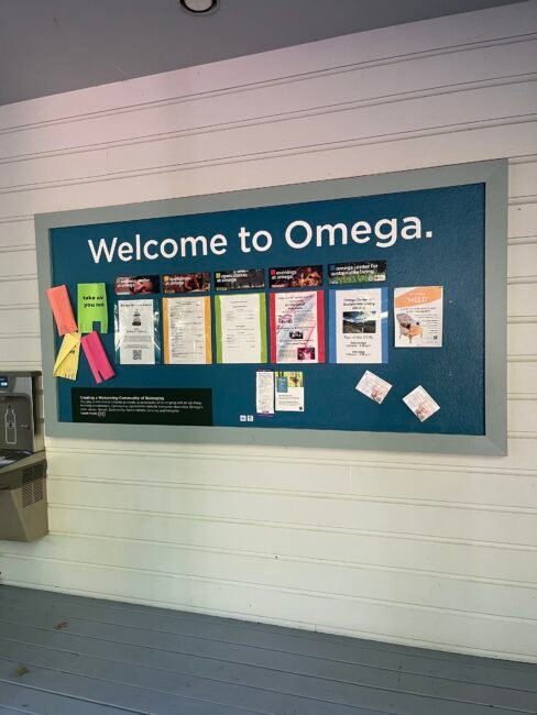 A bulletin board labeled "Welcome to Omega" displays various flyers, notices, and colored paper on a wooden wall near a water fountain.