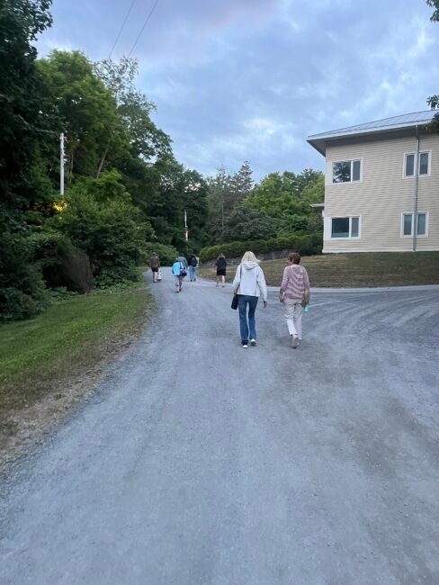 A group of people walk up a wide gravel road bordered by trees and a beige building under a partly cloudy sky.
