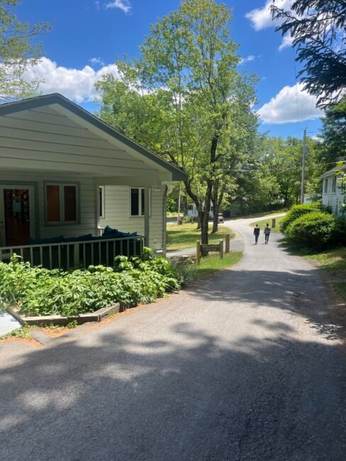 Two people walk down a paved road past a house with a porch and garden, surrounded by trees on a sunny day.