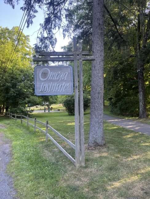 A wooden sign reading "Omega Institute" stands near a gravel path, wooden fence, and green trees on a sunny day.