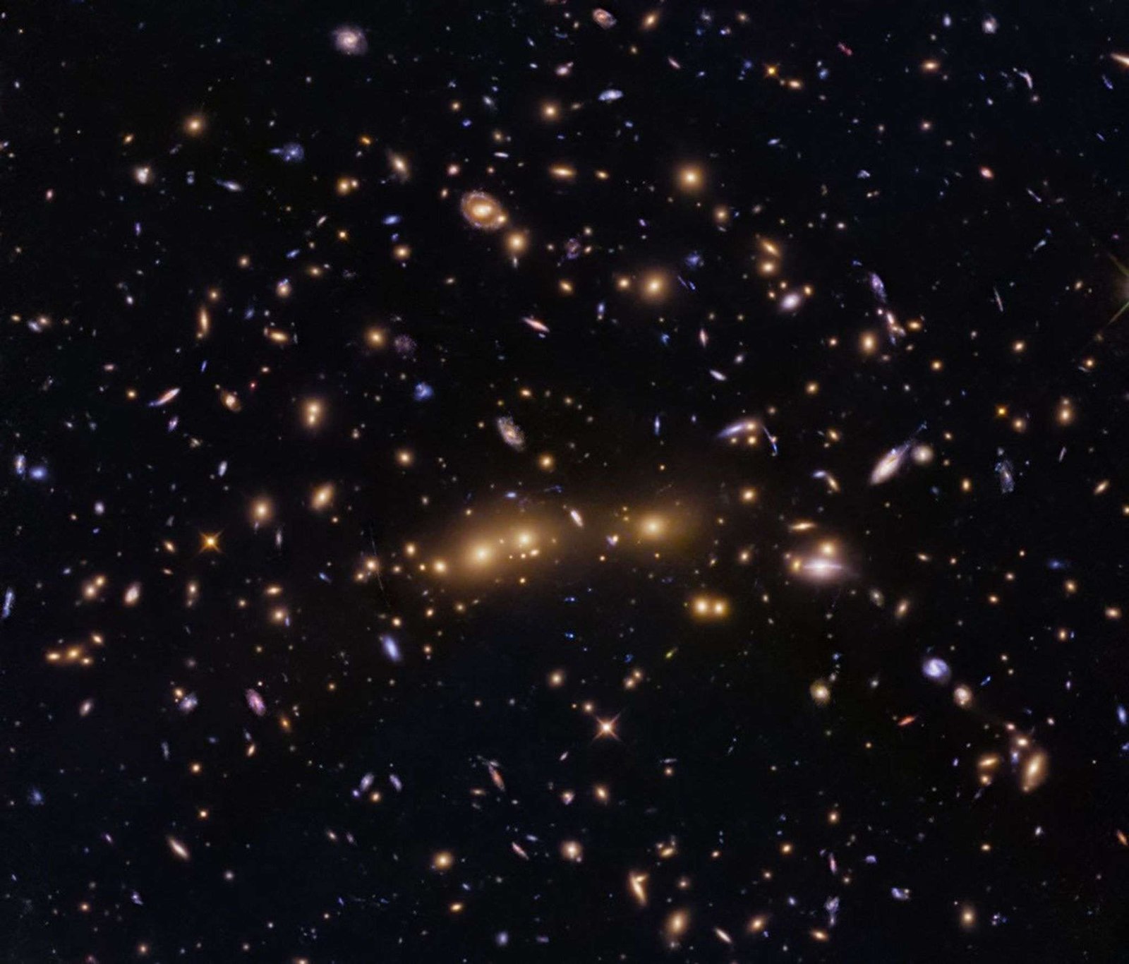 Several massive elliptical galaxies appear to sit in a nearly horizontal line across the center of the image. Numerous spiral, lenticular, and smaller elliptical galaxies dot the space surrounding the giant ellipticals.
