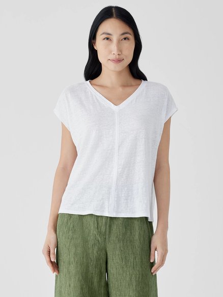 A person with long dark hair stands against a plain background, wearing a white V-neck short-sleeve shirt and green pants.