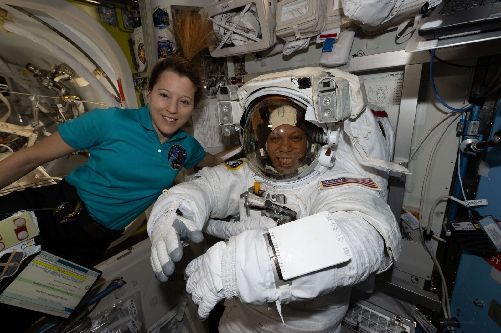 ESA astronaut Sophie Adenot and NASA astronaut Chris Williams smile at the camera. Adenot wears a teal polo shirt with a patch on the front. Williams is in a white spacesuit. They are on the International Space Station.