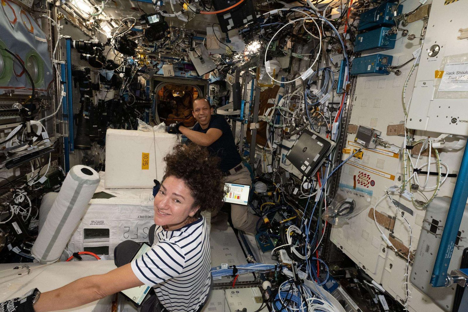 NASA astronauts Jessica Meir is pictured in the foreground wearing a black and white striped t-shirt. Her hair is curly and pulled up at the top of her head. She has a tablet in her lap and mittens on her hands as she faces the camera smiling. NASA astronaut Chris Williams is behind her wearing a black t-shirt and khaki pants, he has a tablet strapped to this upper leg. He is touching a white cube that sits in the middle of the space craft, while looking forward and smiling. The two astronauts are collecting frozen research samples while living and working aboard the International Space Station.