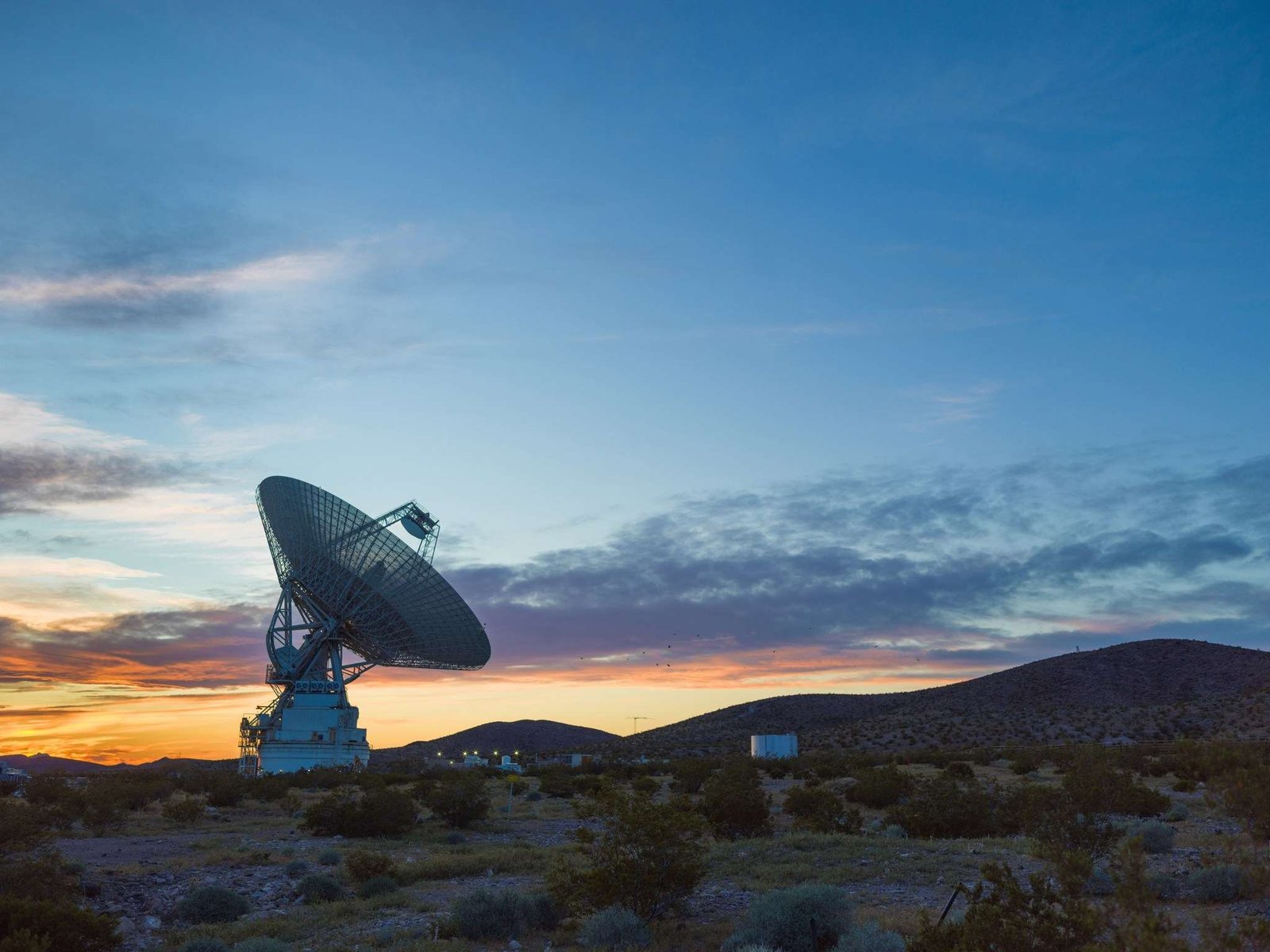 NASA’s Deep Space Station 14, a 230-foot wide radio antenna at the Goldstone Deep Space Communications Complex near Barstow, California is silhouetted against a glowing sunset sky of deep blues, purples, oranges, and yellows. Several support building can also be seen illuminated behind the antenna structure.