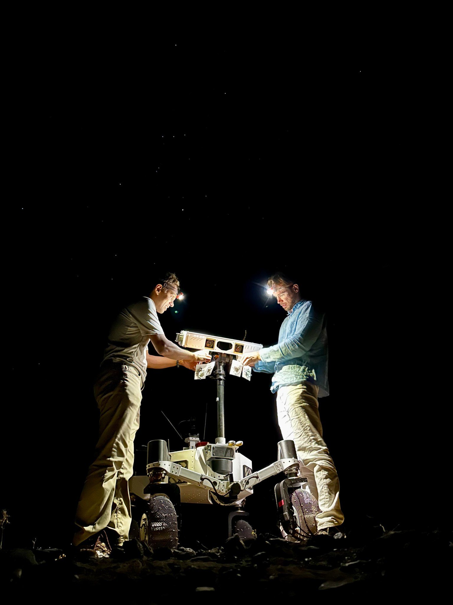 Against a pitch-black background sky in which a handful of white stars are visible, two men stand on either side of a 4.5-foot-tall white rover, which is lit up by the glow from their headlamps as they set up the illuminators on the robot’s mast.