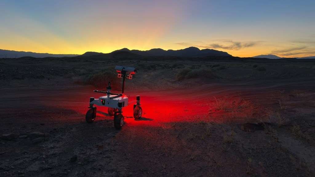 A white four-wheeled rover with a rectangular head on its mast projects a red glow onto a dark desert landscape. Pink and yellow hues indicate the sun has set behind a distant mountain range, while the top of the frame remains blue.
