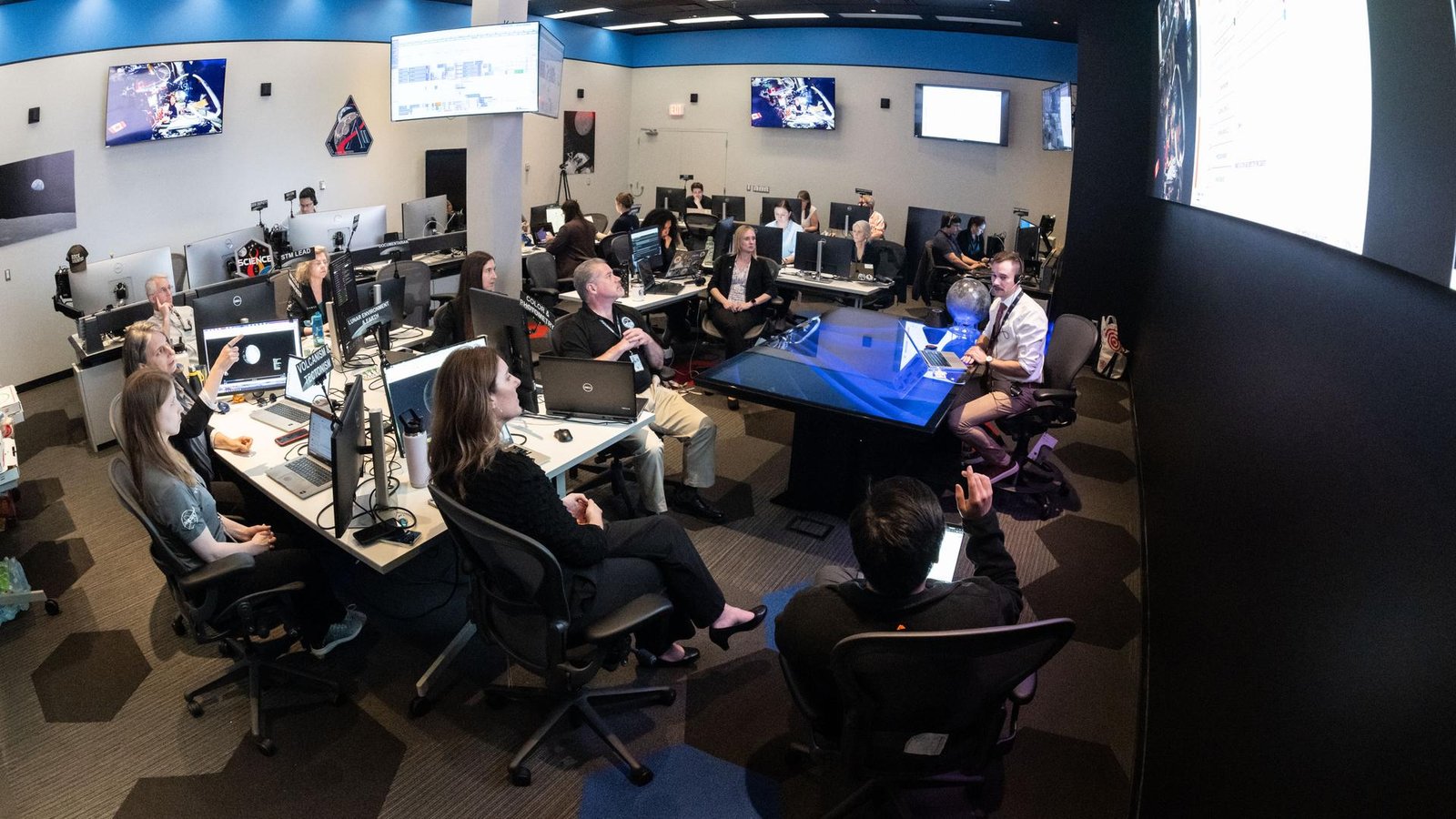 A large operations room filled with about two dozen staff members seated at computer workstations. Rows of desks with multiple monitors face a central area where a person sitting at a glass-topped desk appears to be speaking to the group. Large screens mounted on the walls display space-related imagery and data. Several people are looking towards a large screen hanging on a wall on the right of the image, while others work at their computers. The room is brightly lit, with blue accents along the walls, creating the atmosphere of a space operations or flight control center.