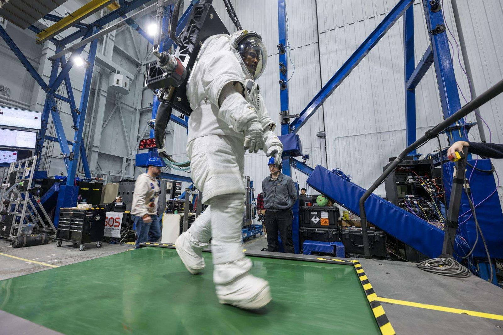 Dressed in a white space suit—with hands gloved, feet booted and head in a helmet with a clear visor—Artemis II astronaut Victor Glover walks on a wide green treadmill. His suit is harnessed to a black machine that is taking the weight off Glover such that, while in the suit, he experiences forces equivalent to lunar gravity. Two people wearing casual clothes and blue hard hats stand in the background, waiting to support Glover and the experiment, should the need arise. Behind Glover are crisscrossing blue iron beams that stretch to a high ceiling in this industrial building at NASA’s Johnson Space Center.