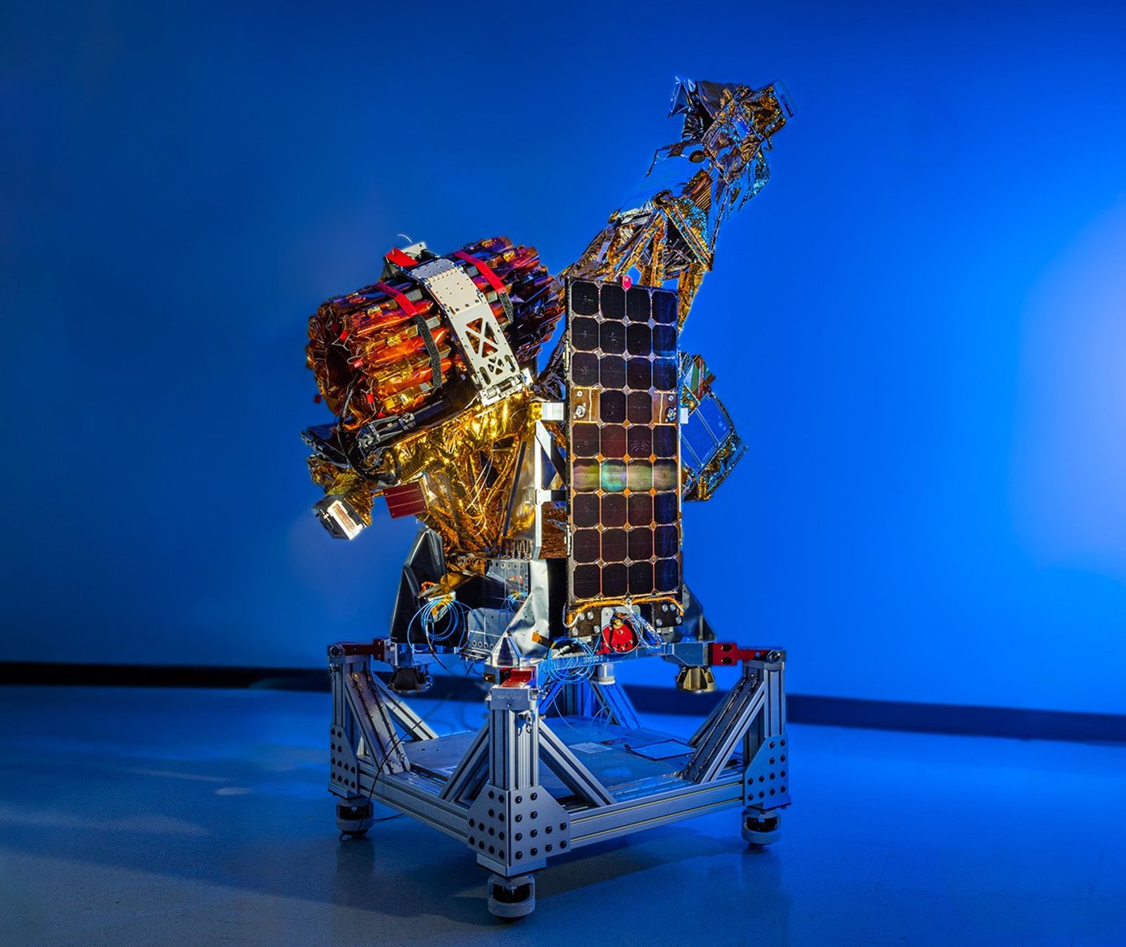 A complex spacecraft partially wrapped in gold-colored foil and featuring a large rectangular solar panel stands on a metallic support structure against a solid blue background.