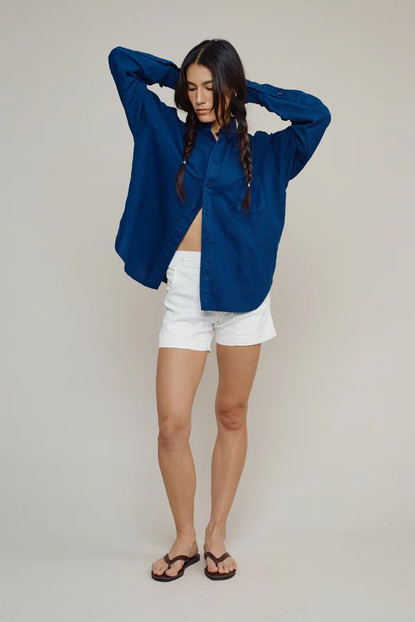 Person standing with arms raised, wearing a loose blue button-up shirt, white shorts, and black flip-flops, against a plain light background.
