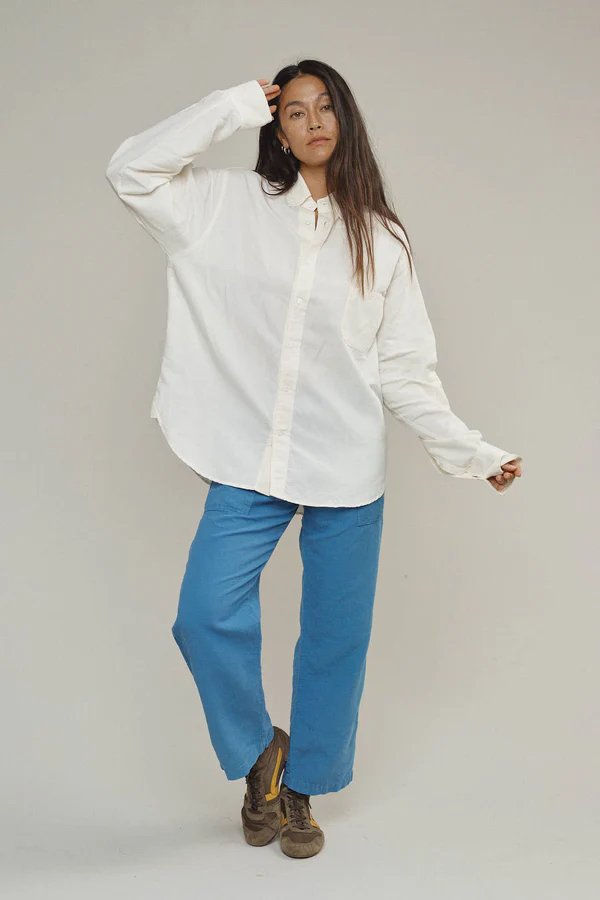 A person stands against a plain background wearing a loose white button-up shirt, blue pants, and brown shoes, with one hand touching their head.