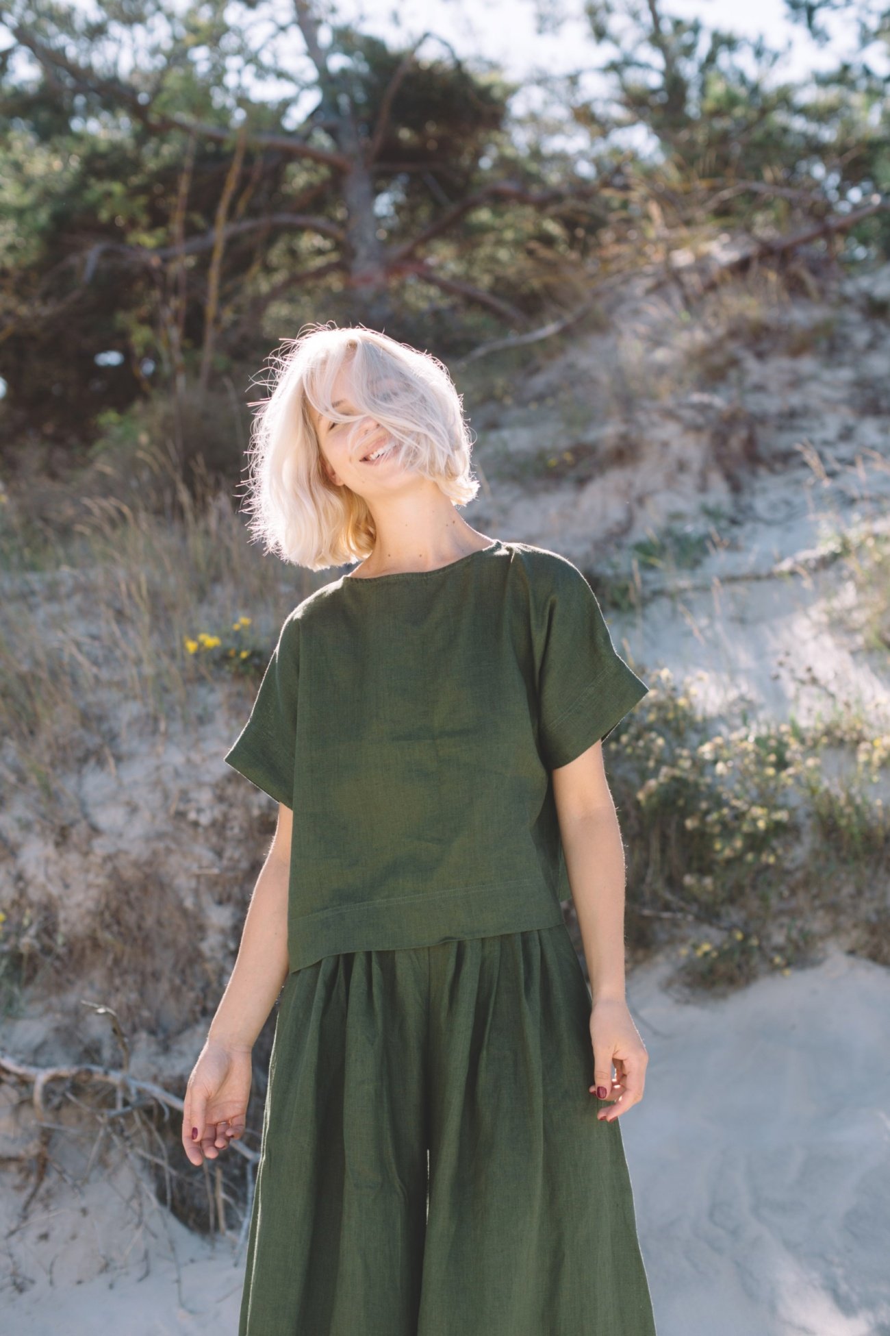 A person with short blonde hair wearing a green top and skirt stands outdoors on sandy ground with vegetation and trees in the background.
