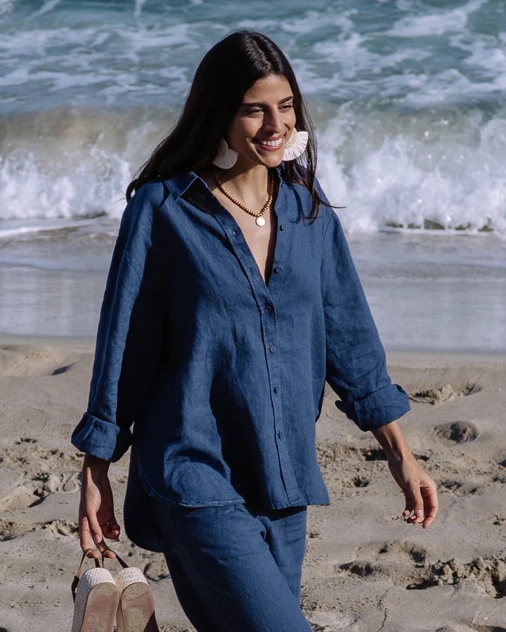 A woman in a blue linen shirt and pants walks barefoot on the beach, holding sandals in one hand, with waves in the background.