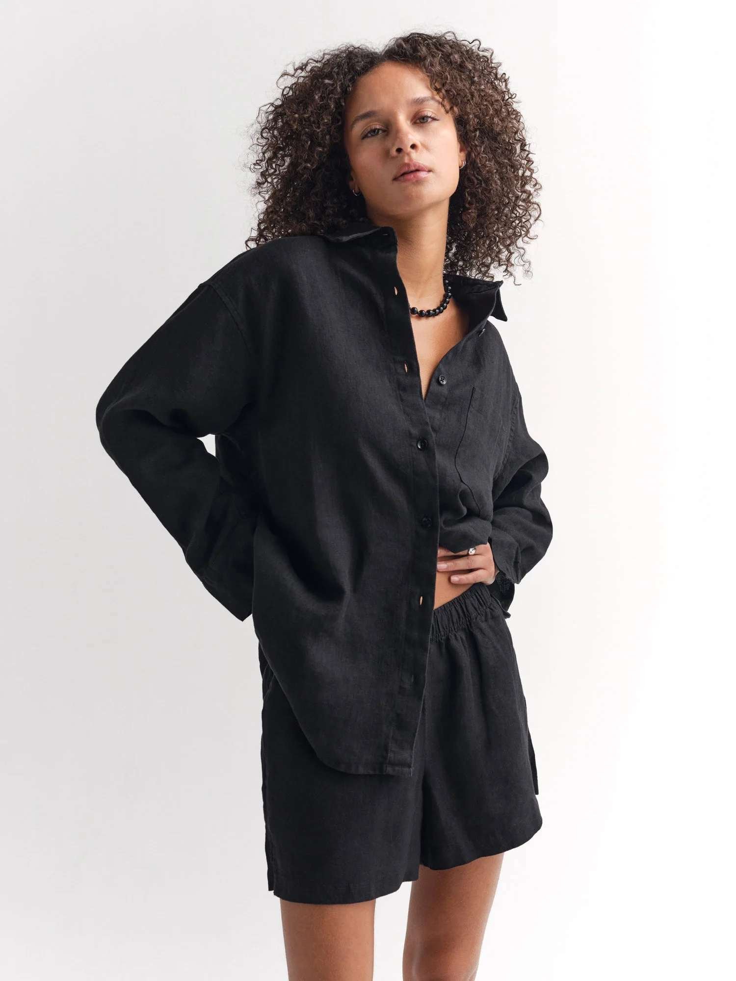A person with curly hair wears a black button-up shirt and matching black shorts, standing against a plain white background.
