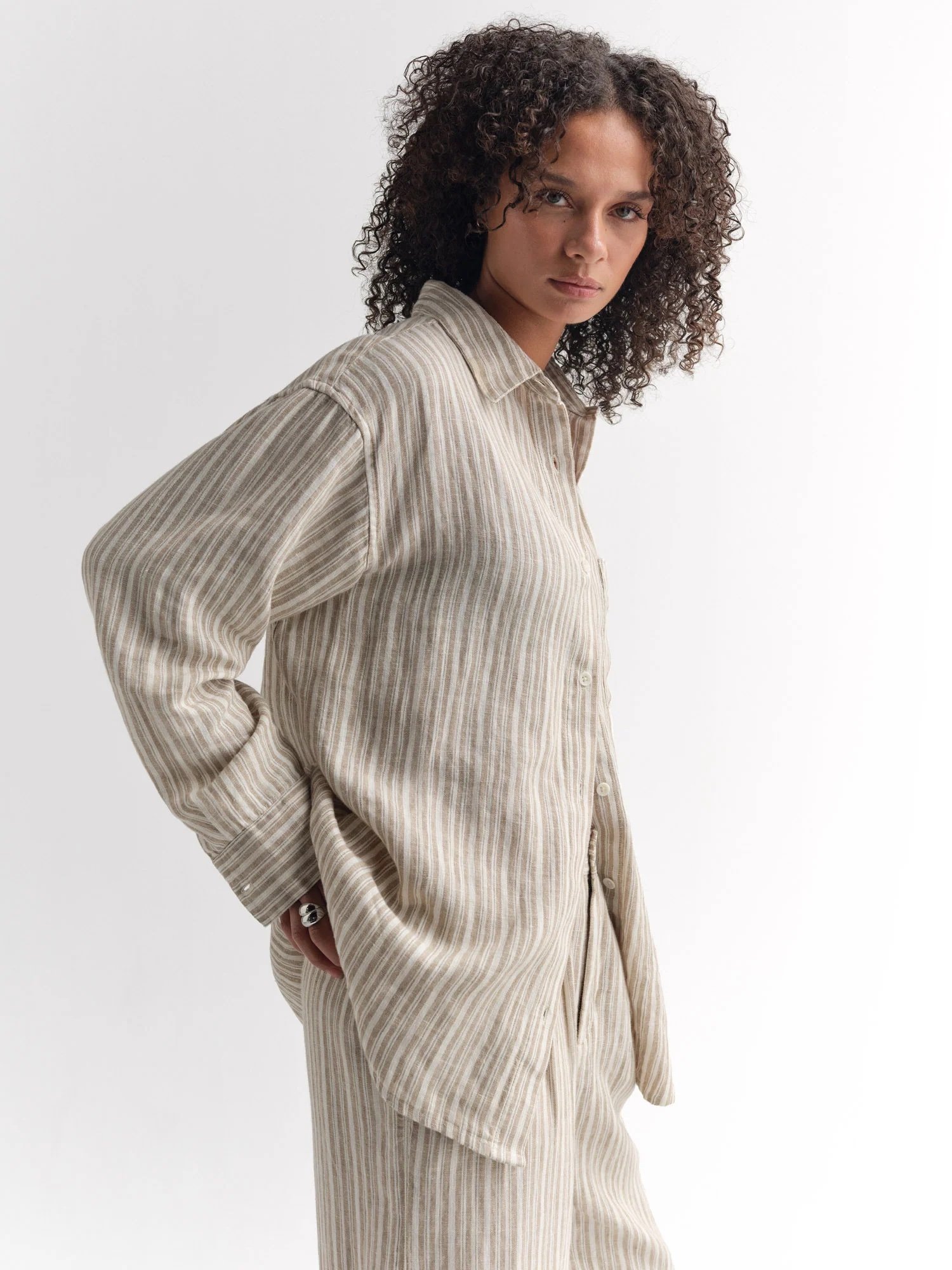 Person with curly hair wearing a loose, beige and white striped button-up shirt and matching pants, standing against a plain white background.