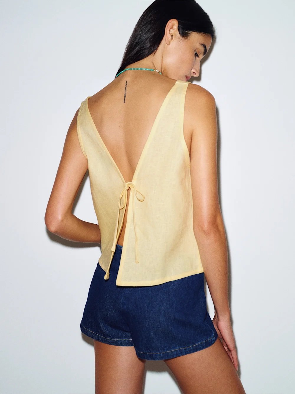 Woman facing backward wearing a sleeveless yellow top with a deep V-back and tie detail, paired with dark denim shorts.
