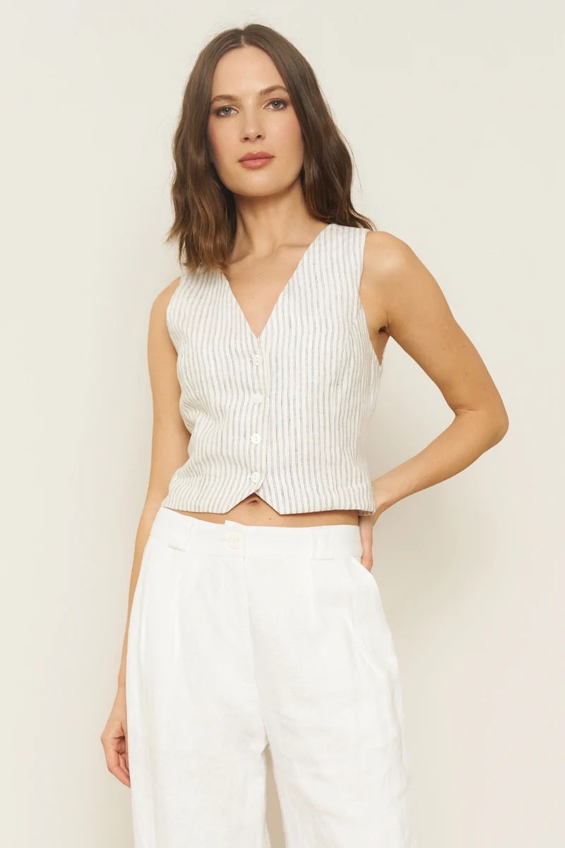 Woman wearing a sleeveless, button-up, striped vest and white pants, standing against a plain light background.