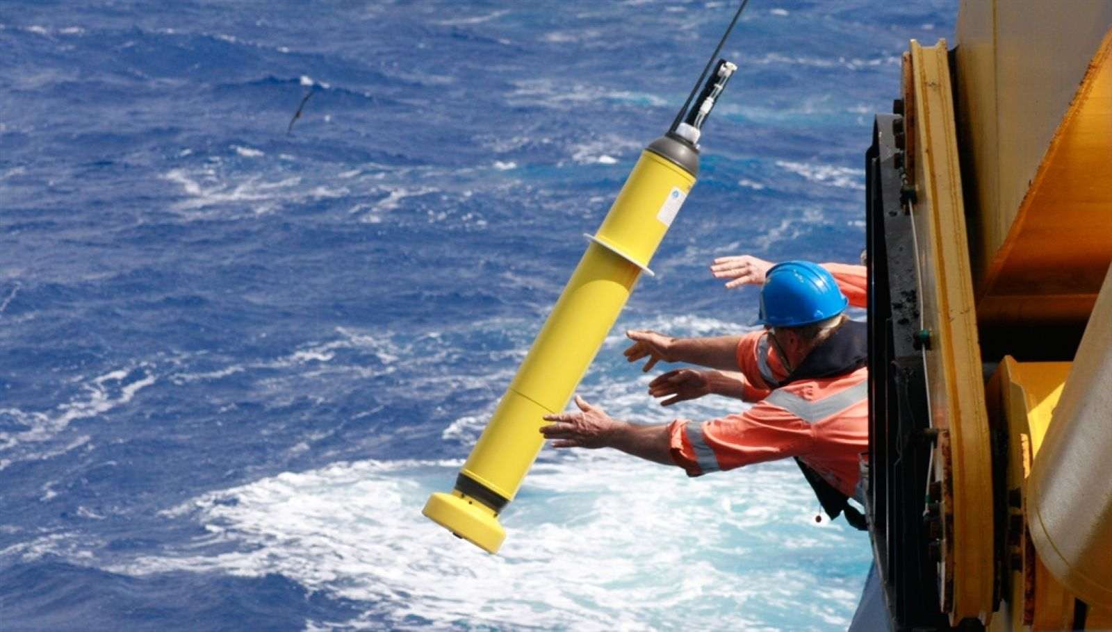a man in an orange shirt tosses a long yellow buoy into the water it is shaped like a pencil but is the size of a large dog
