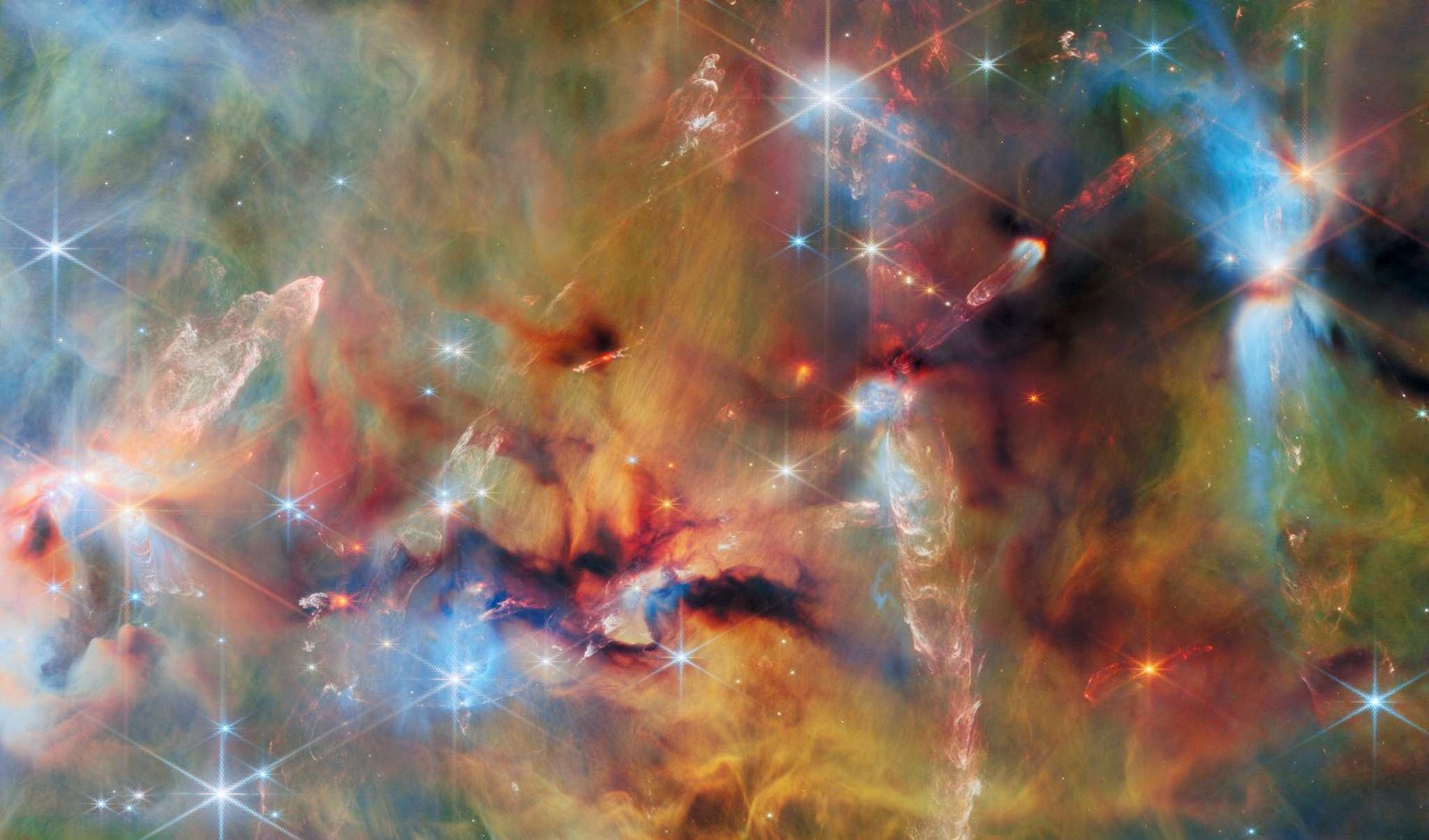 An area inside a star-forming molecular cloud. The background is covered with layers of gas and dust in blue, green and yellowish colors. Thicker clumps of cold dust, dark brown to black, block out light completely. Stars lie among and atop the clouds, from small orange ones to large white or blue ones. Waves and streams of glowing whitish gas are created by jets from protostars colliding with the surrounding material.