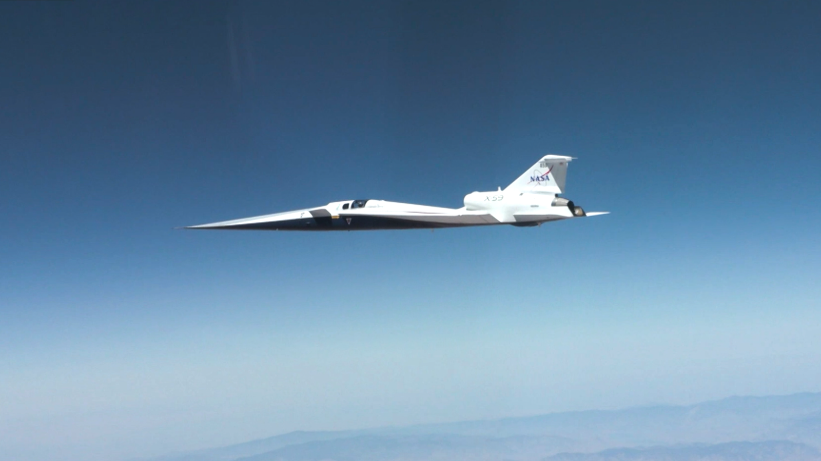 NASA’s X-59 quiet supersonic research aircraft flies through the blue sky. The plane has a distinctive long, sharp nose. There are mountains visible at the bottom of the photo.