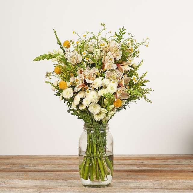 A bouquet of assorted white and yellow flowers in a clear glass jar filled with water, placed on a wooden surface against a plain light background.